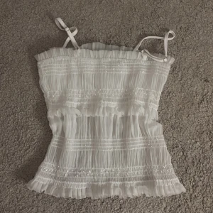 Vit glittrig smockad linnetopp, lite genomskinlig kring midjan men den har ett innerlager vid bysten  - Supersöt vit linnetopp med smockad struktur och tunna axelband. Toppen har glittriga detaljer i tyget och volangkant upptill och nedtill för en romantisk vibe. Perfekt för varma dagar och somriga outfits. Tänk på att den är lite genomskinlig.