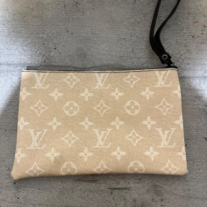 Beige L kuvertväska - Snygg beige kuvertväska L med klassiskt monogrammönster i vitt. Väskan har en avtagbar handledsrem i svart och är tillverkad i tåligt material med exklusiv känsla. Perfekt för dig som vill ha en stilren och ikonisk accessoar.