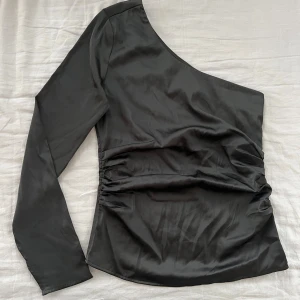 Svart oneshoulder topp från BikBok - Säljer en svart oneshoulder topp från BikBok i glansigt material. Toppen har lång ärm på ena sidan, rynkade detaljer i midjan och dragkedja i sidan för enkel påklädning. 