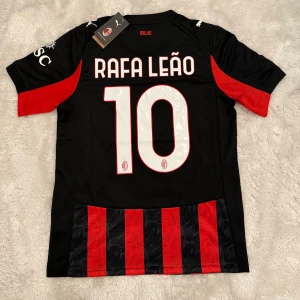 AC Milan Puma fotbollströja Rafa Leão - AC Milan matchtröja från Puma med Rafa Leão och nummer 10 på ryggen. Tröjan är svart och röd med klassiska vertikala ränder, korta ärmar och klubbmärke på bröstet. Tillverkad i lätt och ventilerande material, perfekt för fotbollsplanen.