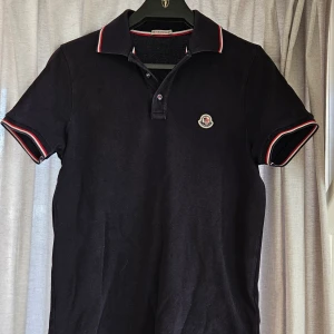 Svart Moncler pikétröja med röd/vit kant - Snygg blå pikétröja från Moncler med klassisk krage och knappar framtill. Tröjan har korta ärmar och detaljer i rött och vitt på krage och ärmslut. Moncler-logga på bröstet och broderade knappar. Perfekt för en stilren och sportig look. Fint skick stl M