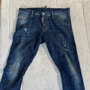 Dsquared2 blå jeans med slitningar - 32/32. Snygga blå jeans från Dsquared2 med coola slitningar och färgstänk på benen. Klassisk femficksmodell med normal passform och låg midja. Jeansen har en mörk tvätt och detaljerade sömmar, perfekt för dig som gillar en edgy streetstil.