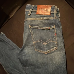 Replay Anbass blå jeans, stl 29 - Snygga Replay Anbass jeans i klassisk blå tvätt. Modellen har raka ben, fem fickor och detaljerade sömmar på bakfickan. Skinnpatch med Replay-logga baktill och röd broderad detalj. Perfekt passform och stilren look.
