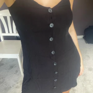 Svart kort klänning med knappar - Säljer en svart kort klänning med smala axelband och knappar hela vägen framtill. Klänningen har en enkel och stilren design med lätt figurnära passform. 