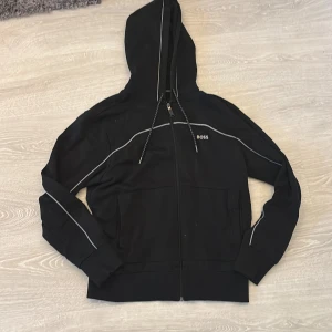 Svart hoodie från BOSS med dragkedja - Svart hoodie från BOSS med vit piping längs axlar och ärmar, samt broderad BOSS-logga på bröstet. Tröjan har huva med snörning, hel dragkedja och två fickor framtill. Materialet är mjuk bomullsmix som känns skön mot huden.