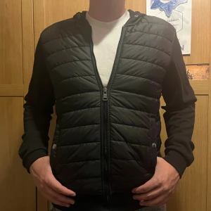 Svart Moncler pufferjacka med huva - Säljer en svart äkta pufferjacka från Moncler i storlek S. Jackan har quiltad front, huva och Moncler-logga på ärmen. Dragkedja framtill och två fickor med knapp. Perfekt för dig som vill ha en stilren och sportig look.