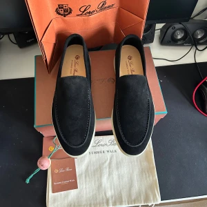 Svarta mockaloafers Loro Piana Summer Walk - Stilrena svarta loafers från Loro Piana, modellen Summer Walk. Skorna är i mjuk mocka med vit platt sula och diskret logga bak. Perfekta för en clean och lyxig look, med klassisk rund tå och minimalistisk design.