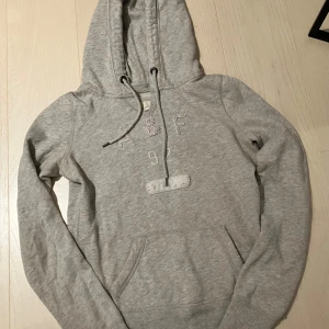 Grå hoodie Abercrombie & Fitch XS - Grå hoodie från Abercrombie & Fitch i mjuk bomull med broderad A&F-logga och siffran 92 framtill. Lila paljettdetaljer på bokstäverna, stor huva med snörning och klassisk känguruficka. 