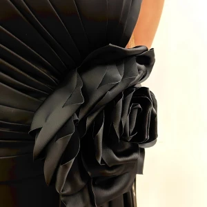 Svart långklänning med blomdetalj - Superelegant svart långklänning från Mavina med en stor, dramatisk blomdetalj i midjan och snygga draperingar. Klänningen har en hög slits på sidan och är tillverkad i ett glansigt, följsamt material som ger en lyxig känsla.