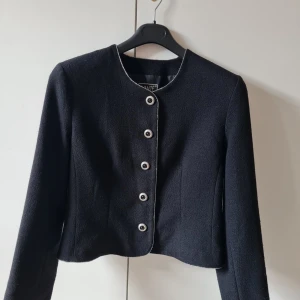 Kavaj från Avante strl 42 - Stilren svart kavaj från Avante med rund halsringning och dekorativa knappar framtill. Jackan är croppad och har långa ärmar med knappar vid ärmsluten. Storlek 42. Kan hämtas i Vallentuna.