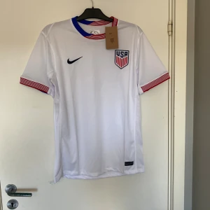 USA fotbollströja Nike Dri-FIT L - Vit officiell USA fotbollströja från Nike i Dri-FIT-material. Tröjan har korta ärmar med röda och vita ränder vid ärmslut och hals, samt blå detalj i kragen. Broderad USA-logga på bröstet och Nike-logga. Perfekt för dig som vill representera USA på planen.