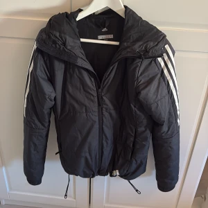 Svart pufferjacka från Adidas S - Svart pufferjacka från Adidas med huva och klassiska vita ränder på ärmarna. Jackan har dragkedja framtill, snörning i nederkant och långärmade ärmar. Perfekt för kyliga dagar och har en sportig vibe med snygg passform.
