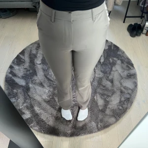 Beige kostymbyxor - Snygga beige kostymbyxor med hög midja. Byxorna har klassiska fickor fram och bak, samt hällor för bälte. Stängs med dold hake, knapp och dragkedja. Materialet är slätt och känns dressat, perfekt för en clean look.