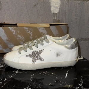 Golden Goose sneakers med glitterstjärna - Snygga vita sneakers från Golden Goose med silverfärgade detaljer och en glittrig stjärna på sidan. Skorna har grå snörning, rund tå och platt sula. Ovandelen är i skinn och hälen har metallic-silver finish. Perfekta för dig som gillar statement-detaljer.