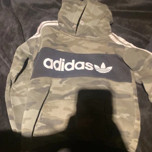 Camouflage hoodie från Adidas - Säljer en grön camouflage hoodie från Adidas med stor vit logga framtill och klassiska ränder på ärmarna. Tröjan har huva, lång ärm och är tillverkad i mjukt material. Perfekt för dig som gillar streetwear och sportig stil.
