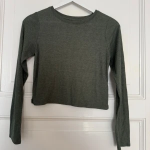 Ribbad grön långärmad topp H&M  - En ribbad långärmad topp från H&M i en dämpad grön nyans. Modellen är croppad med rund halsringning och smal passform. Perfekt att styla med jeans eller kjol för en enkel och snygg look.