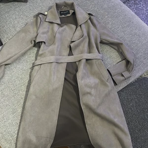 Beige trenchcoat i mockaimitation Missay XS - Snygg beige trenchcoat från Missay i mockaimitation. Jackan har klassisk krage, bälte i midjan och långa ärmar. Perfekt för lager-på-lager och ger en clean look till alla outfits. Fickor på sidorna och axelklaffar för extra detalj.