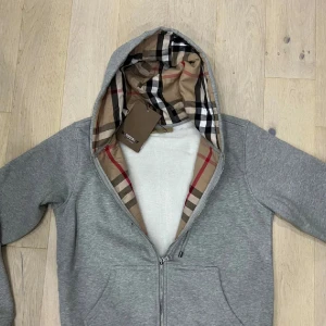 Grå Burberry hoodie med rutig huva - Grå hoodie från Burberry med dragkedja och klassisk rutig insida i huvan. Tröjan har snörning vid huvan, broderad Burberry-logga på bröstet och två fickor fram. Tillverkad i mjuk bomull, perfekt för chill dagar. Snyggt ribbade muddar och normal passform.