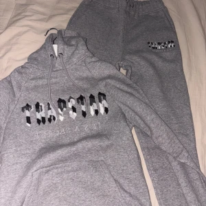 Trapstar dress - Säljer en grå hoodie från Trapstar med svart, vit och gråt mönstrat tryck på bröstet och texten 'IT'S A SECRET' under. Hoodien har snörning i huvan, magficka och långa ärmar. Perfekt för chill dagar och streetwear-stil.