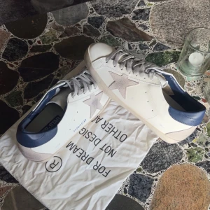 Golden Goose sneakers med blå detalj - Snygga Golden Goose sneakers i vitt skinn med grå mockastjärna på sidan, grå snörning och mörkblå hälkappa. Klassisk låg modell med platt sula och rund tå. Perfekta för dig som gillar stilrena men ändå unika sneakers.