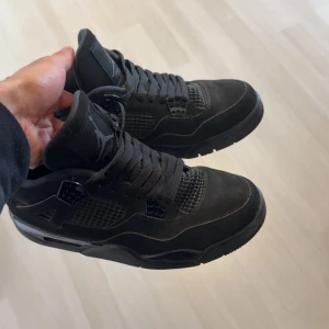 Svarta Nike Air Jordan 4 sneakers - Säljer ett par helsvarta Nike Air Jordan 4 sneakers med klassisk siluett, meshdetaljer och Jumpman-logga på plös och häl. Skorna har snörning, robust sula och är tillverkade i mocka och syntet. Perfekta för dig som gillar streetstyle och vill sticka ut med en ikonisk modell.