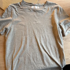 Grå t-shirt från Les Deux - Snygg grå t-shirt från Les Deux med enkel design och liten vit logga på bröstet. Klassisk rund halsringning och korta ärmar. Perfekt basic-plagg i mjuk bomull som passar till allt.