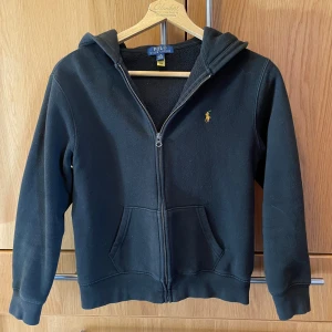 Svart hoodie från Polo Ralph Lauren - Svart hoodie från Polo Ralph Lauren med dragkedja och klassisk gul broderad logga på bröstet. Tröjan har huva, känguruficka och är tillverkad i mjukt material som passar perfekt för chill dagar. Enkel och stilren design som funkar till det mesta. Sitter som xs i herrstorlek! Pris kan diskuteras.