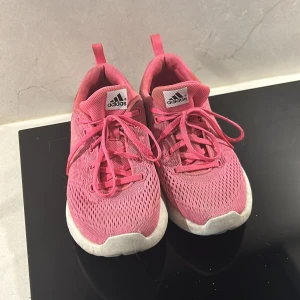 Rosa löparskor från Adidas - Säljer ett par rosa Adidas löparskor och vita sulor. Skorna har snörning och en sportig, lätt design som passar perfekt till vardags eller träning. Adidas-logga på plösen. Snygga och bekväma för dig som gillar färg!