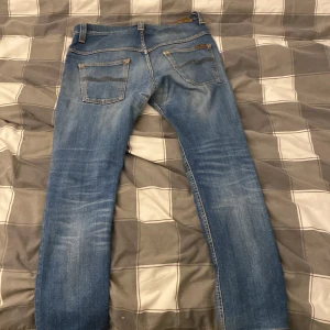 Blå jeans från Nudie Jeans Co - Snygga blå jeans från Nudie Jeans Co med klassisk femficksdesign och orangea sömmar. Jeansen har en rak passform och subtila slitningar framtill och baktill. Nudie-logga på bakfickan och läderpatch med logotyp i midjan. Perfekt för dig som gillar stilrena jeans. har en liten defekt som inte syns 