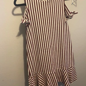 Randig kortklänning med knyt i ärmarna - Superfin kortklänning från Zara med röda och vita ränder. Klänningen är ärmlös med snygga knyt-detaljer på axlarna och har en volang nertill. Dragkedja i ryggen för enkel påklädning. Perfekt för dig som gillar en lekfull och trendig stil.