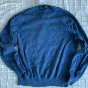 Blå stickad tröja från Polo Ralph Lauren - Klassisk blå stickad tröja från Polo Ralph Lauren med rund halsringning och broderad logga på bröstet. Tröjan har ribbade muddar vid ärmslut och nederkant, perfekt för lager-på-lager under höst och vinter.