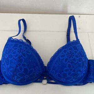 Blå spets bh  med blommönster - Snygg blå bh med blommigt spetsmönster och justerbara axelband. Kuporna är vadderade och har dekorativ ringdetalj framtill. Baksidan har klassisk hysk- och hakknäppning. Perfekt för dig som vill ha något extra under kläderna.