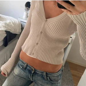 Beige ribbad kofta - Säljer en beige ribbad kofta med knappar framtill. Den är croppad och har en v-ringad design som ger en stilren look. Perfekt för att matcha med jeans eller kjol. Tror att storleken är M men passar small( S)