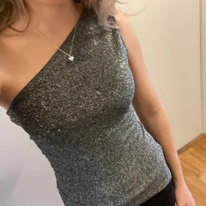 Glittrig one shoulder top - Säljer en glittrig silverfärgad topp från H&M med one shoulder-design. Toppen är tight och har en snygg glansig yta som verkligen sticker ut. Omsydda från L till S och passar perfekt till nyår eller jul🩷