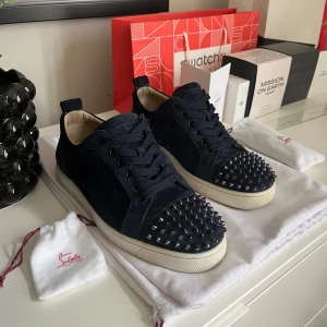 Christian Louboutin sneakers med nitar - Snygga mörkblå sneakers från Christian Louboutin i mocka med ikonisk röd sula och svarta nitar på tån. Skorna har snörning och vit platt sula, perfekt för dig som vill sticka ut med en edgy och lyxig stil. Storlek 40 passar 41