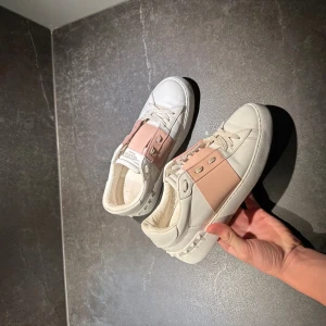 Valentino Garavani sneakers vit/rosa - Säljer ett par snygga Valentino Garavani sneakers i vitt skinn med bred rosa rem och coola nitar på sidorna. Skorna har snörning och en chunky vit sula med rutmönster undertill. Perfekta för dig som vill ha en lyxig och trendig look.