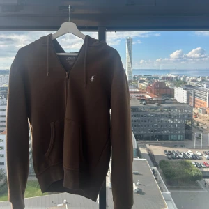 Brun hoodie från Polo Ralph Lauren - Säljer en brun hoodie från Polo Ralph Lauren med dragkedja och huva. Tröjan har en klassisk känguruficka framtill och den ikoniska vita loggan broderad på bröstet. Perfekt för chill dagar och snygg till jeans eller joggers. Pris 2395kr i butik