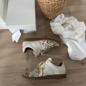 Maison Margiela Gats ”Paintsplatter”🎨 - Unika Maison Margiela gats sneakers i beige skinn med coola färgstänk i gult, rött, blått och svart. Klassisk låg modell med snörning och rund tå. Snygg statement-sko för dig som gillar konstnärlig vibe och streetstyle. Box, dustbags, kvitto medföljer och skorna är givetvis äkta!