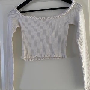 Vit ribbad croppad topp FB Sister - Superfin vit ribbad croppad topp från FB Sister med långa ärmar och volangdetaljer vid halsringning och nederkant. Toppen har en offshoulder-design och är perfekt för dig som gillar en söt och trendig stil.