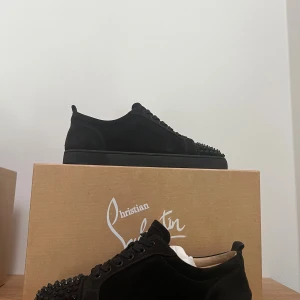 Christian Louboutin svarta sneakers med nitar - Svarta sneakers från Christian Louboutin i mocka med ikoniska röda sulor och svarta nitar på tån. Skorna har snörning och en platt sula, vilket ger en edgy och lyxig vibe. Perfekta för dig som vill sticka ut med exklusiva detaljer.