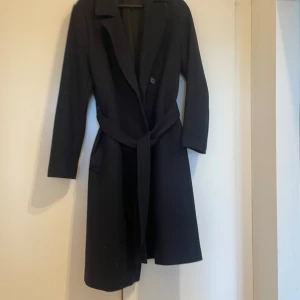 Svart kappa från Filippa K med bälte - Stilren svart kappa från Filippa K i storlek medium. Klassisk design med lång ärm, krage och knäppning framtill. Kappan har ett matchande bälte i midjan för snygg passform och är tillverkad i mjukt ullmaterial. Perfekt för dig som gillar minimalistisk och tidlös stil.