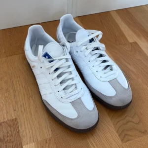 Adidas Samba OG vita sneakers - Säljer ett par klassiska Adidas Samba sneakers i vitt skinn med grå mockadetaljer och bruna gummisulor. Skorna har de ikoniska tre ränderna på sidan, blå logga på plösen och snörning framtill. Perfekta för dig som gillar retrostil och streetwear. Har använts 2 gånger och är impregnerade. Säljes pga brist på plats.