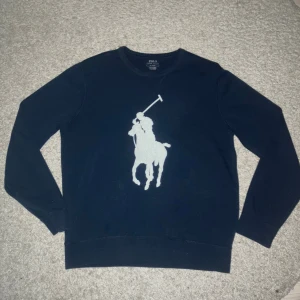 Mörkblå tröja från Polo Ralph Lauren - Snygg mörkblå tröja från Polo Ralph Lauren med stor vit polospelare på bröstet. Klassisk rund halsringning och långa ärmar. Tillverkad i mjuk bomull som är skön att ha på sig. Perfekt för dig som gillar stilrena och sportiga plagg.