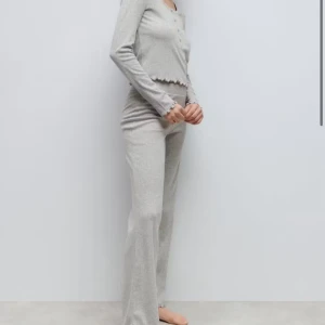 Grå ribbade mjukisbyxor med volangkant + matchande tröja  - Supermysiga grå mjukisbyxor med ribbad struktur och volangkant längst ner. Byxorna har en skön loose passform och är perfekta för chill dagar hemma. Materialet är mjukt och stretchigt, vilket gör dem extra bekväma. Medföljer även matchande tröja, så hela settet!