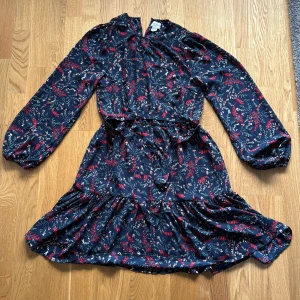 Blommig kortklänning Holly & Whyte - Supersöt marinblå kortklänning från Holly & Whyte med blommigt mönster i rött och vitt. Klänningen har långa ärmar, volangkant nertill och knytband i midjan. Perfekt för dig som gillar en feminin och lekfull stil.