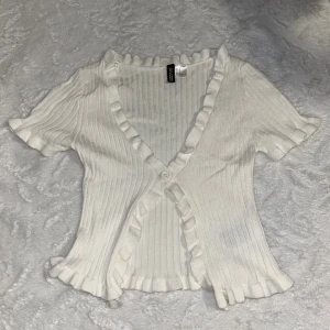 Vit ribbad blus med volang från H&M Divided - Supersöt vit ribbad blus från H&M Divided med korta ärmar, volangkant längs v-ringningen och nederkanten. Blusen har en knapp framtill och är croppad. Superfint på 🥰