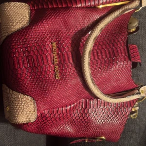 Röd handväska Michael Kors i skinn - Säljer en snygg röd handväska från Michael Kors med ormskinnsmönster och detaljer i beige. Väskan har guldfärgade metalldetaljer, innerfack med dragkedja och klassisk MK-logga. Perfekt för dig som vill sticka ut med en unik accessoar.