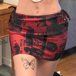 Röd och svart kortkjol med tryck - Säljer en röd och svart kortkjol med unikt tryck kjolen har snörning i sidorna för justerbar passform och är figurnära. 