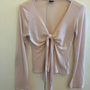 Ljusrosa topp från Gina Tricot - Supersöt ljusrosa topp från Gina Tricot med lång arm och knyt framtill. Toppen har en djup v-ringning och är tillverkad i mjuk bomullsmix. Perfekt att styla med jeans eller kjol för en trendig look.