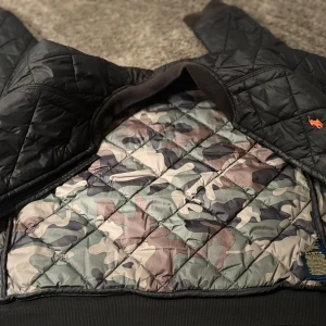 Svart quiltad jacka Polo Ralph Lauren - Quiltad svart jacka från Polo Ralph Lauren med orange logga på bröstet. Insidan har ett snyggt camouflagemönster i dova färger. Jackan är tillverkad i polyester och har ribbade muddar nertill. Perfekt för höst och vinter.